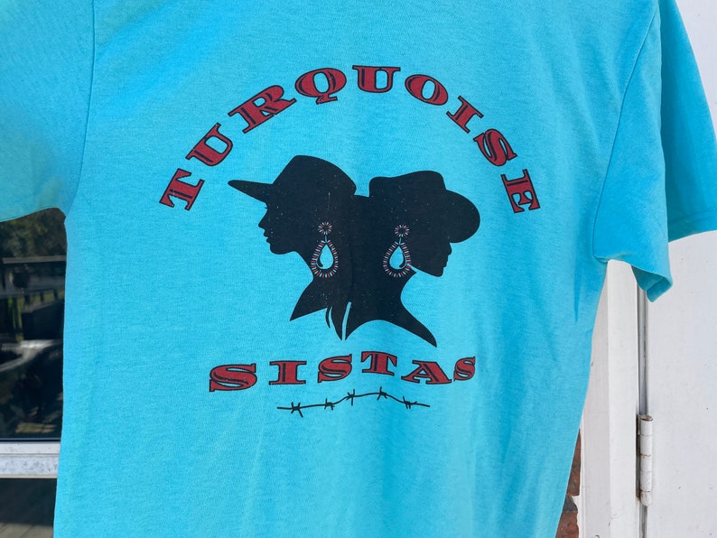 May include: A turquoise blue t-shirt featuring a black silhouette of two women wearing cowboy hats and earrings. The text "TURQUOISE SISTAS" is printed in red on the shirt.