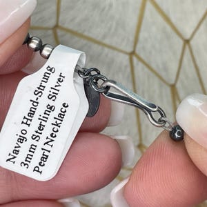 Peut inclure: Gros plan sur un collier de perles Navajo Hand-Strung en argent sterling de 3 mm. Le collier est compos&eacute; de petites perles gris fonc&eacute; enfil&eacute;es sur un fil fin, avec un fermoir argent&eacute;. Une &eacute;tiquette blanche avec la description du collier est attach&eacute;e.