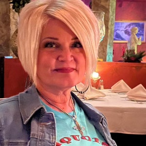 May include: A woman with blonde hair is wearing a blue denim jacket over a turquoise t-shirt with a red and black graphic that says "Cowgirl Sisters".
