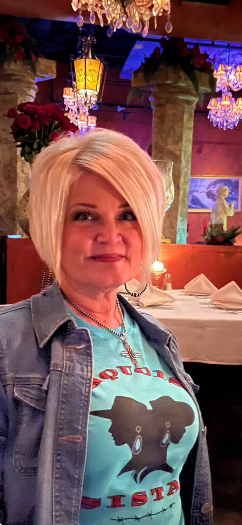 May include: A woman with blonde hair is wearing a blue denim jacket over a turquoise t-shirt with a red and black graphic that says "Cowgirl Sisters".