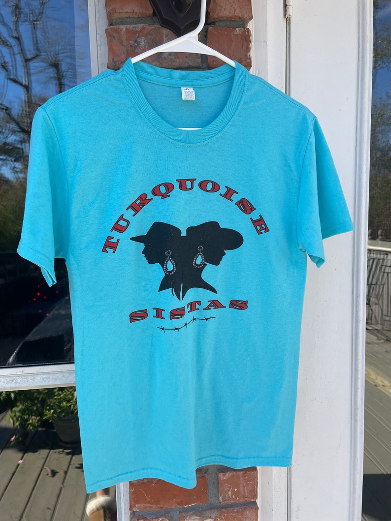 May include: A turquoise blue t-shirt with a red and black graphic of two women wearing cowboy hats and the text "TURQUOISE SISTAS".