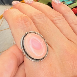May include: A silver ring featuring a large, oval-shaped pink gemstone. The gemstone has a lighter pink center and is set in a silver bezel with decorative details. The ring is a statement piece, likely for fashion or jewelry.