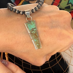 May include: A silver pendant necklace with a turquoise and brown stone inlay. The pendant is rectangular with a geometric design and is suspended from a beaded necklace.