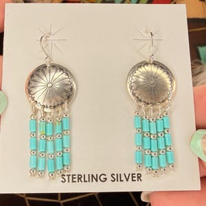 May include: A pair of sterling silver dangle earrings featuring a circular stamped metal disc at the top. Each earring has four hanging strands composed of alternating small silver beads and cylindrical turquoise-colored beads.