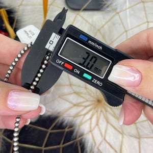 Peut inclure: Gros plan sur un collier de perles argent&eacute;es mesur&eacute; avec un pied &agrave; coulisse num&eacute;rique. Le pied &agrave; coulisse affiche une mesure de 3,0 mm. Une &eacute;tiquette avec du texte est visible. En arri&egrave;re-plan, un attrape-r&ecirc;ves.