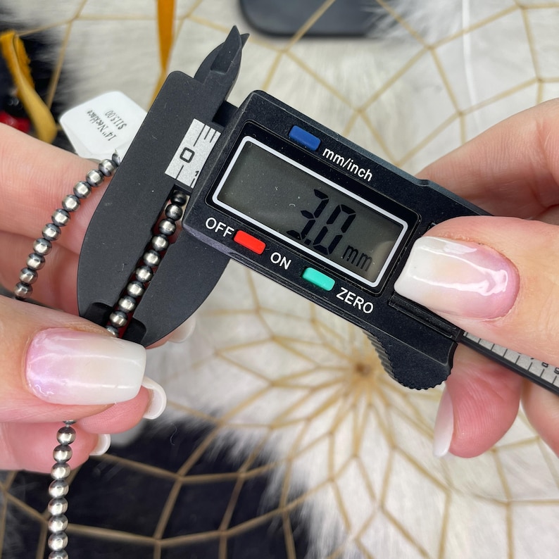 Peut inclure: Gros plan sur un collier de perles argent&eacute;es mesur&eacute; avec un pied &agrave; coulisse num&eacute;rique. Le pied &agrave; coulisse affiche une mesure de 3,0 mm. Une &eacute;tiquette avec du texte est visible. En arri&egrave;re-plan, un attrape-r&ecirc;ves.