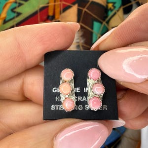 May include: A pair of sterling silver earrings with three pink and peach-colored stones. The earrings are held on a black card with text that reads "GENUINE HAND CRAFTED STERLING SILVER."