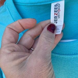 May include: A light blue t-shirt with a white tag that says "JERZEES" and "DRI-POWER" in black text. The tag also says "S" for size, "Cotton" and "Polyester" for fabric content, and "Made in El Salvador" for origin.