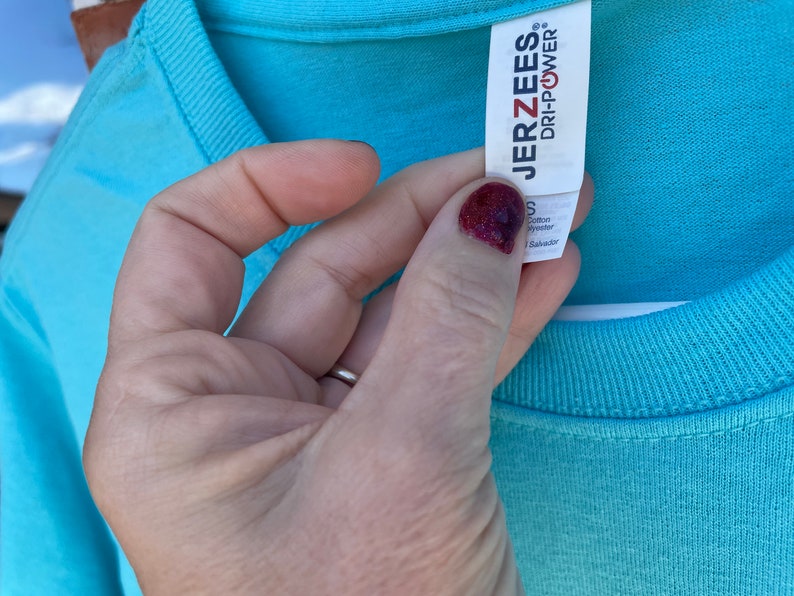 May include: A light blue t-shirt with a white tag that says "JERZEES" and "DRI-POWER" in black text. The tag also says "S" for size, "Cotton" and "Polyester" for fabric content, and "Made in El Salvador" for origin.
