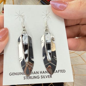 May include: A pair of sterling silver dangle earrings shaped like feathers. Each earring features a central inlay of black and white stone segments. The earrings are attached to a white display card with the text GENUINE HANDCRAFTED STERLING SILVER.