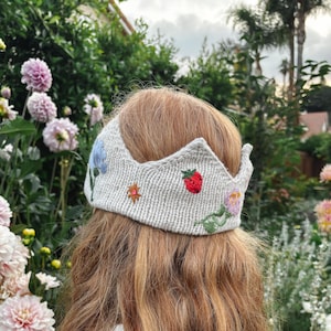 May include: A knitted crown with embroidered flowers and a strawberry. The crown is light beige and has a scalloped edge. The flowers are blue, pink, and orange. The crown is worn on a person's head.