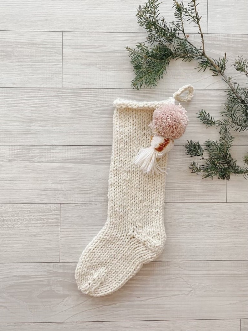 May include: A white knitted Christmas stocking with a pink and white pom-pom on top. The stocking is hanging on a white wooden background with a green pine branch.