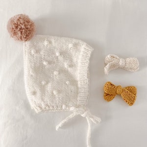Può includere: Un berretto per neonato in maglia bianca con un pon pon marrone in cima. Il berretto ha un laccio in basso ed è decorato con piccole palline bianche. Sono inclusi anche due fiocchi in maglia, uno bianco e uno giallo senape.