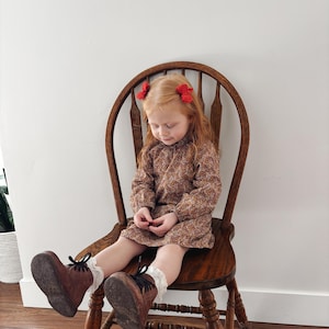 May include: A young girl wearing a brown floral dress and brown boots sits on a wooden chair. She has red hair with red bows in her hair.