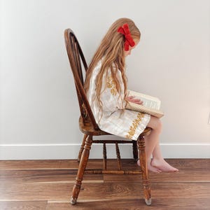 May include: A young person with long hair wearing a red bow hair clip, seated on a wooden chair, reading a book. The person is wearing a white dress with gold embroidery. The chair is made of dark wood and has a spindle back.