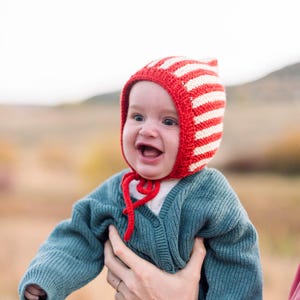 May include: A baby wearing a red and white striped knitted hat with a blue cardigan and blue jeans.