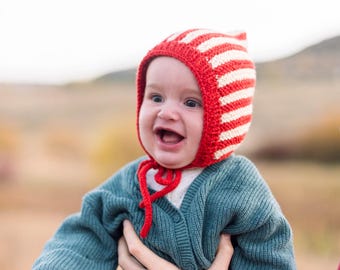 Puede incluir: Un bebé con un gorro de punto rojo y blanco a rayas, un cárdigan azul y unos vaqueros azules.