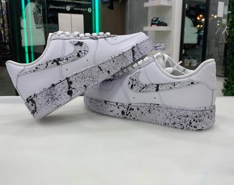 May include: A pair of white sneakers with black paint splatters. The sneakers have a white swoosh logo on the side and a white sole.
