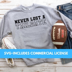 May include: A gray sweatshirt with the text "NEVER LOST A TAILGATE" printed on the front. A brown football with white laces is lying on the sweatshirt. The sweatshirt is on a white wooden surface.