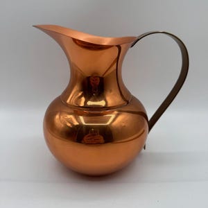 May include: A copper-colored pitcher with a curved handle. The pitcher has a bulbous body and a flared spout. The handle is a darker, contrasting color. The surface of the pitcher is highly reflective, showing the surrounding environment.