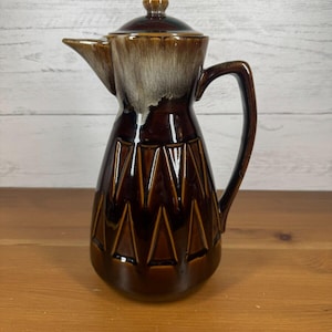 May include: A brown ceramic coffee pot with a geometric pattern and a lid. The pot has a white glaze on the top and a brown glaze on the bottom.