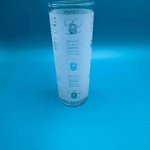 Puede incluir: Un vaso alto y transparente con un diseño esmerilado. El diseño presenta un ratón de dibujos animados y el texto "Tim Horton BEIGNES". El vaso tiene una forma ligeramente acampanada y está sobre un fondo azul.