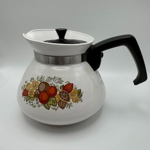 Könnte beinhalten: Weiße Emaille-Kaffeekanne mit schwarzem Griff und Deckel. Die Kanne hat ein dekoratives Blumendesign mit orangefarbenen, braunen und gelben Blumen und Früchten. Ein silbernes Band umschließt die Kanne in der Nähe der Oberseite.