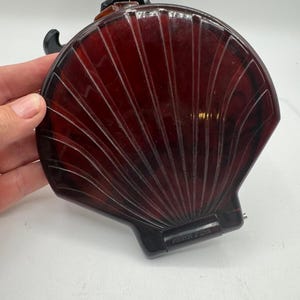 Puede incluir: Un espejo compacto con forma de concha en color burdeos oscuro, con acabado brillante y estrías radiales moldeadas. El estuche con bisagra presenta una silueta curva y suave, con un pequeño mecanismo de bisagra metálica en la base.