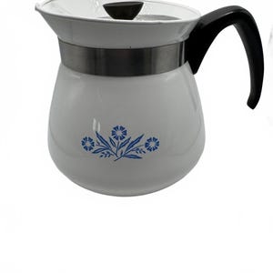 Könnte beinhalten: Eine weiße Emaille-Teekanne mit einem blauen Blumenmuster auf der Vorderseite. Die Teekanne hat einen schwarzen Griff, einen passenden schwarzen Deckelknopf und ein silberfarbenes Metallband, das den oberen Teil des Körpers unter dem Deckelrand umschließt.