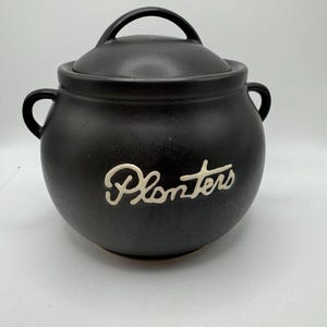 Puede incluir: Tarro de mantequilla de cacahuete Planters de cerámica negra con tapa y dos asas. El tarro tiene la palabra "Planters" escrita en blanco en la parte delantera.