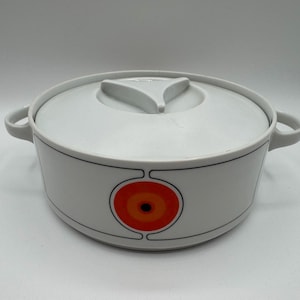 May include: White ceramic casserole dish with a lid and handles. The dish has a black trim and a circular design with orange and yellow concentric circles. The lid has a triangular handle.