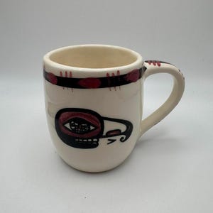 Può includere: Tazza in ceramica color crema con un design nero e rosso. La tazza presenta un volto stilizzato sul davanti e una fascia di punti rossi e linee nere attorno al bordo. Il manico è anch'esso color crema.