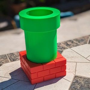 Puede incluir: Una tubería de plástico verde se encuentra sobre una estructura de ladrillos rojos. La tubería tiene un borde ancho y un cuerpo cilíndrico. La estructura de ladrillos está hecha de bloques rectangulares apilados. Los objetos probablemente están inspirados en un videojuego.