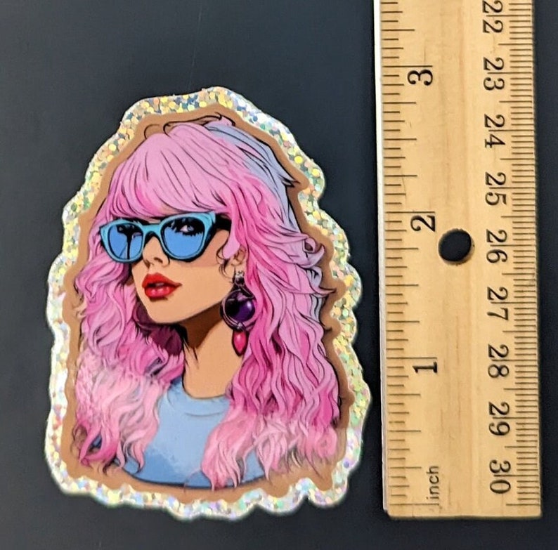May include: A glittery sticker featuring a cartoon illustration of a woman with pink hair, blue sunglasses, and a blue shirt. The sticker is approximately 2.5 inches tall.
