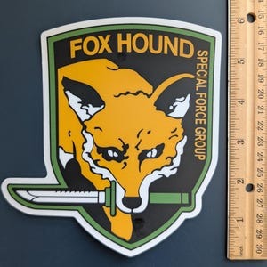 Puede incluir: Adhesivo en forma de escudo con una cabeza de zorro naranja estilizada, detalles en blanco y negro y un cuchillo con mango verde. El texto "FOX HOUND SPECIAL FORCE GROUP" está en amarillo y blanco. El adhesivo mide unos 7.6 cm de alto.