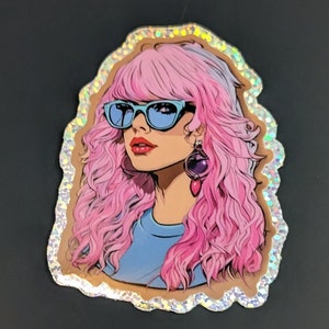 May include: A sticker featuring a cartoon illustration of a woman with pink hair, wearing blue sunglasses and a blue shirt. The sticker has a glittery, iridescent border.
