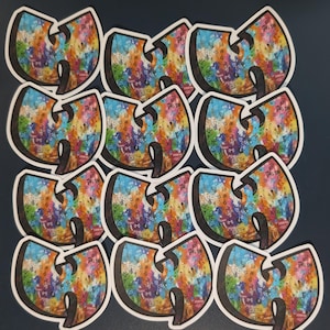 Puede incluir: Una colección de pegatinas con el logotipo de Wu-Tang Clan de colores. Cada pegatina presenta un diseño abstracto vibrante con un contorno negro y un borde blanco. Las pegatinas están dispuestas sobre una superficie oscura.