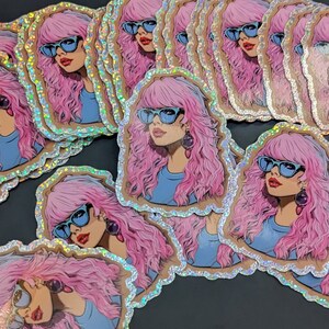 May include: A stack of holographic stickers featuring a woman with pink hair wearing blue sunglasses. The stickers have a glittery, iridescent finish.