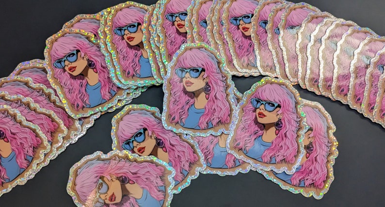 May include: A stack of holographic stickers featuring a woman with pink hair wearing blue sunglasses. The stickers have a glittery, iridescent finish.