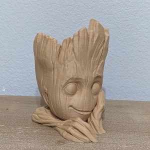 May include: A light brown, 3D-printed planter shaped like the character Groot from Guardians of the Galaxy. The planter features Groot's face and arms, with a textured wood-like appearance. The head is open to hold plants.