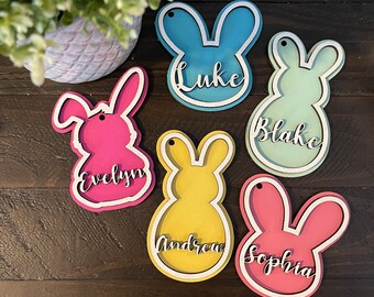 May include: A collection of colorful wooden bunny-shaped name tags. Each tag features a different color, including pink, blue, yellow, and green, with a white outline. Names like "Luke," "Evelyn," and "Sophia" are displayed in cursive.