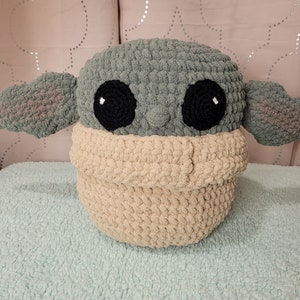 Puede incluir: Un juguete de peluche tejido a crochet con forma de personaje de la franquicia Star Wars. El juguete es verde y marrón con ojos negros y un acento blanco en cada ojo. El juguete está sentado sobre una superficie azul claro y peluda.
