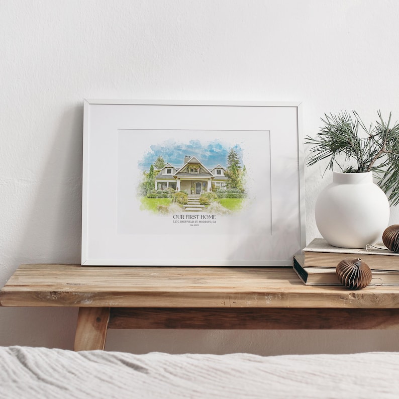 May include: A white framed print with a watercolor illustration of a house. The text "OUR FIRST HOME" is below the image, along with the address and date.