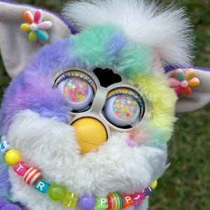 Puede incluir: Un juguete Furby colorido con un cuerpo de arcoíris, pelaje blanco y ojos llenos de estrellas. Tiene un collar de cuentas que deletrea "TRIPP" y pendientes con forma de flor. Los colores del juguete incluyen morado, azul, verde, amarillo y rosa.