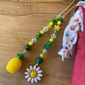 May include: Two beaded hair accessories. One features a yellow pineapple charm, the other a white and yellow daisy. Both have yellow, green, and clear beads. The accessories are on a wooden surface, near a doll wearing a red dress.