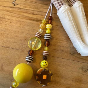 May include: Two beaded charms with yellow, brown, and white beads. One charm has a large yellow sphere, the other has a pancake charm with a smiley face. White boots and fishnet stockings are in the background.