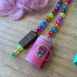 May include: A pink My Melody tumbler with a handle and a cartoon graphic, paired with a beaded charm necklace featuring colorful beads. A brown domino charm is also attached to the necklace. The background includes pink hair.
