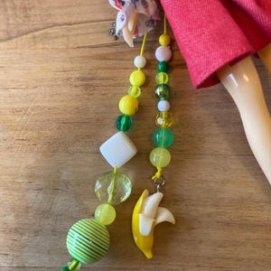 May include: Two beaded charms with yellow cords. One charm features a yellow banana pendant. The other charm has a variety of beads in shades of yellow, green, and white, including a striped ball and a clear, faceted bead.
