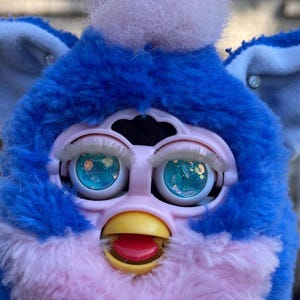 May include: A close-up of a vintage Furby toy. The toy has blue fur, light pink accents, and large, expressive eyes with glittery irises. It features a yellow beak and a red tongue, with white eyelashes.