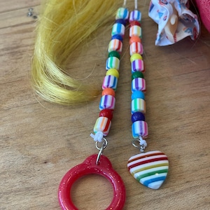 May include: Close-up of a doll's hair with colorful beaded accessories. The accessories include a red ring and a heart-shaped charm with rainbow stripes. The beads are in various colors, including blue, green, yellow, and orange.